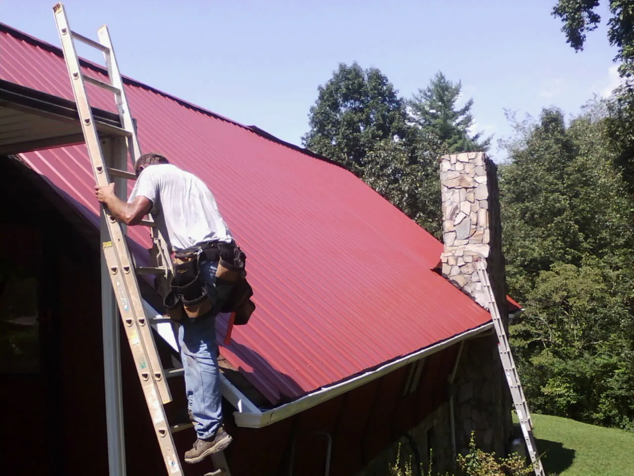 Roofer on a ladder beside a red metal roof and stone chimney — Soffit & Fascia Repair by Roofing Brothers in South Gardiner